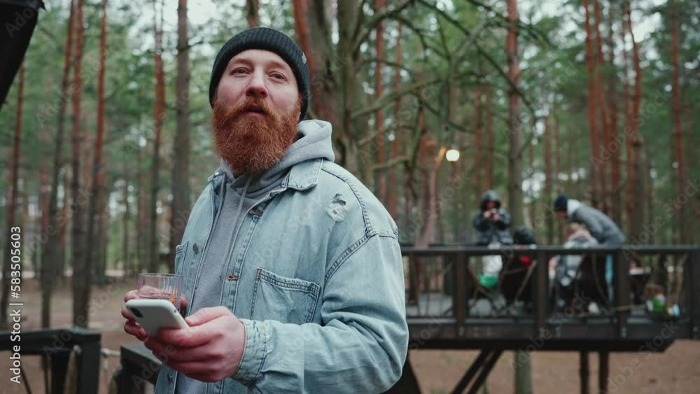 Smiling bearded tourist man texting on mobile and looking around with friends around the campfire in the forest Smiling bearded tourist man texting on mobile and looking around with friends around the campfire in the forest