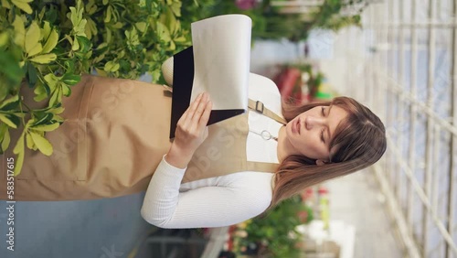 Side view of female florist satnding, working in greenhouse, inspecting plants. Beautiful, focused lady holding folder, taking notes. Concept of growing and taking care of plants. photo