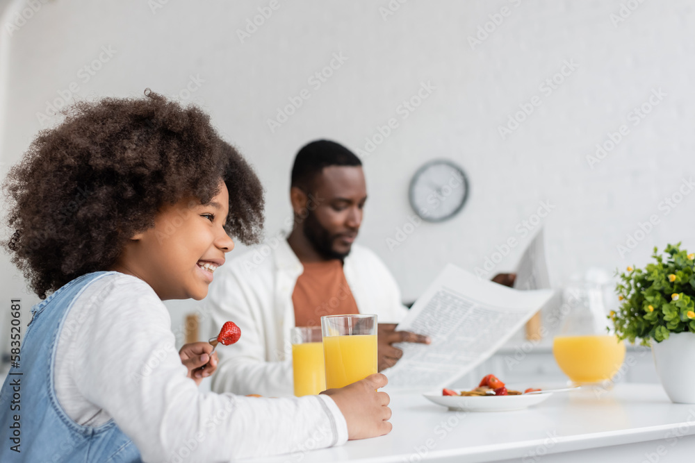 happy african american girl having breakfast while father reading newspaper on blurred background. happy african american girl having breakfast while father reading newspaper on blurred background.