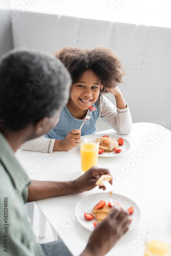 happy african american girl looking at grandfather eating pancakes during breakfast.