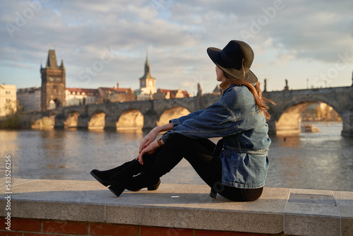 Stylish beautiful young woman wearing black hat sitting on Vltava river shore in Prague with Charles Bridge on background. Elegant retro lady fine art portrait.