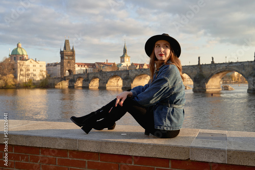 Stylish beautiful young woman wearing black hat sitting on Vltava river shore in Prague with Charles Bridge on background. Elegant retro lady fine art portrait.