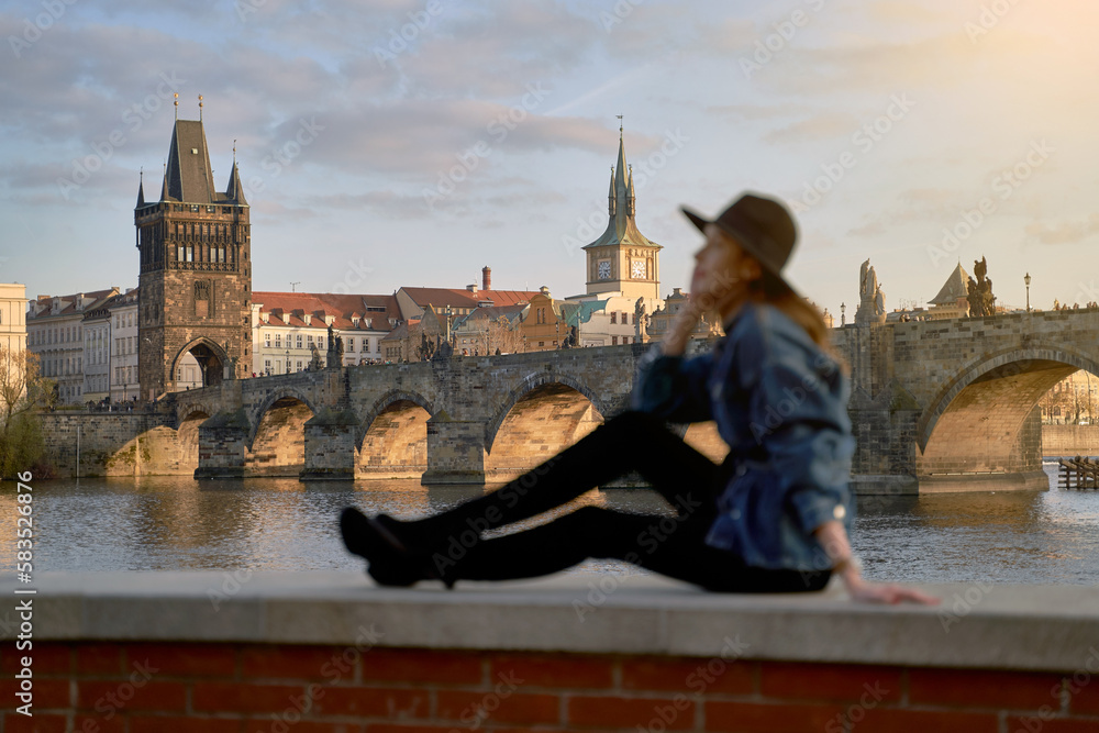 Stylish beautiful young woman wearing black hat sitting on Vltava river shore in Prague with Charles Bridge on background. Focus on background.