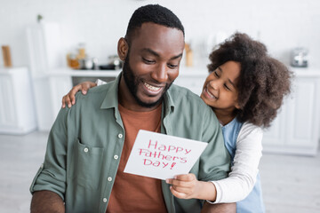 african american girl giving greeting card with happy fathers day lettering to cheerful dad.