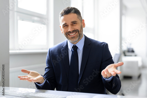Successful middle aged businessman sitting at desk in office, gesturing and smiling at camera
