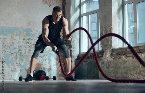 Strength and endurance. Young muscular man doing workout, training with battle ropes indoors. Functional training exercises. Concept of sportive lifestyle, body care, fitness, hobby, health