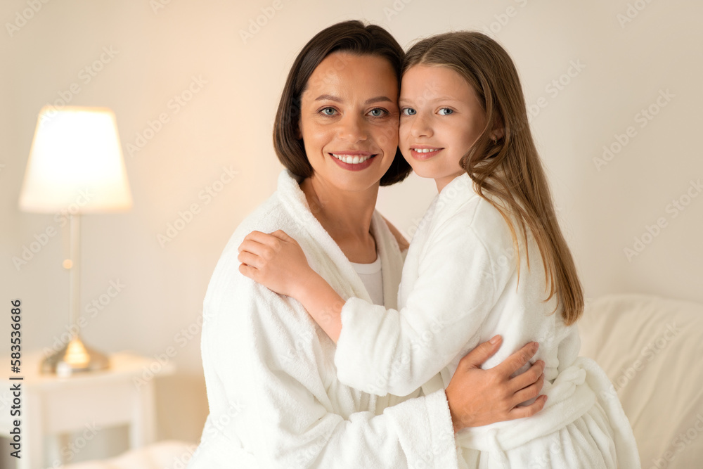 Smiling european millennial lady and little girl in bathrobes hugs on bed, enjoy rest together, free time and weekend Smiling european millennial lady and little girl in bathrobes hugs on bed, enjoy rest together, free time and weekend