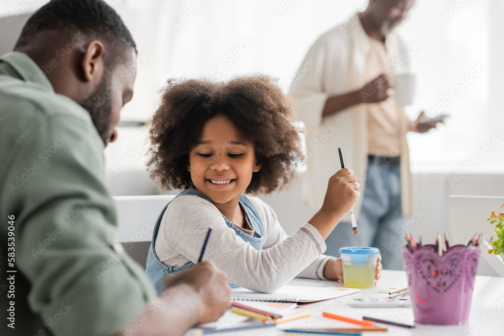 Cheerful african american girl painting with blurred dad near grandparent at home.