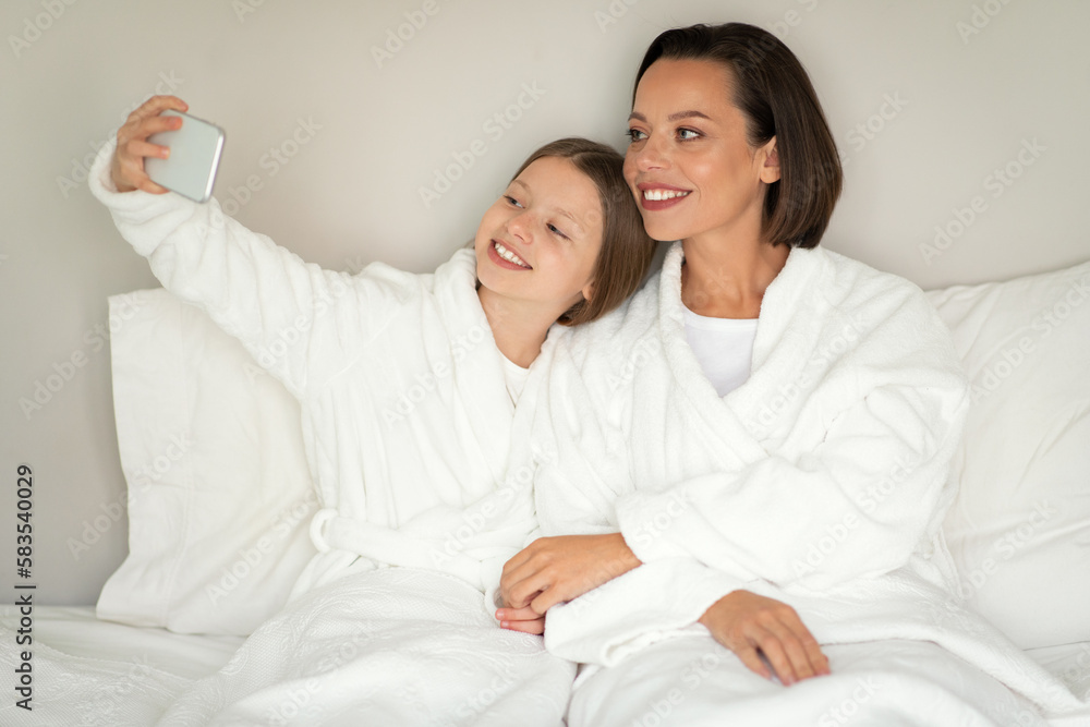 Smiling young female, little caucasian girl in bathrobe take selfie on phone, shoot video for blog, relax Smiling young female, little caucasian girl in bathrobe take selfie on phone, shoot video for blog, relax