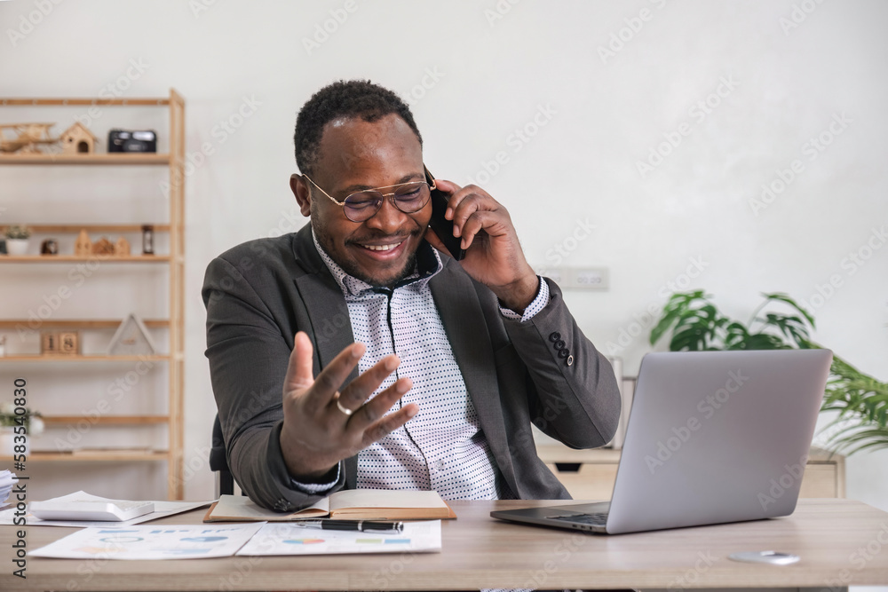Happy black businessman talking on cellphone having phone conversation sitting at workplace working on laptop computer in office. Happy black businessman talking on cellphone having phone conversation sitting at workplace working on laptop computer in office.