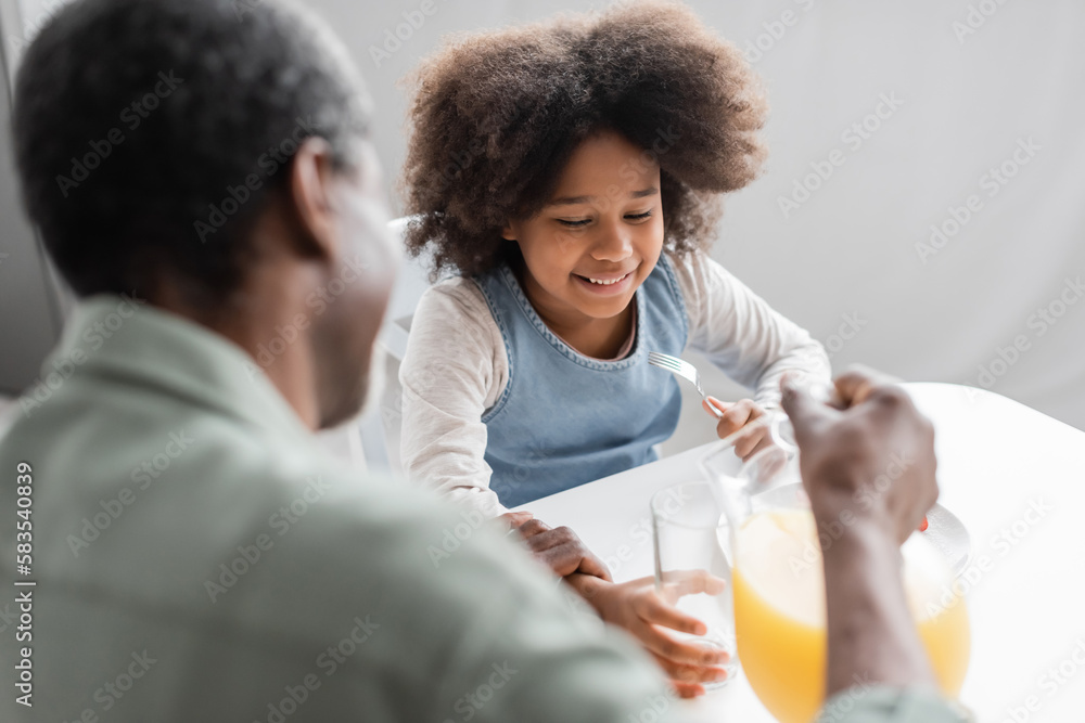 happy african american girl holding fork while grandpa pouring orange juice during breakfast. happy african american girl holding fork while grandpa pouring orange juice during breakfast.