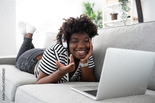 Happy young African American woman having video call on laptop while lying down on sofa. Black woman broadcasting or live streaming. Online communication concept