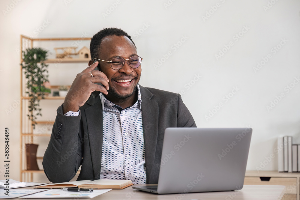 Happy black businessman talking on cellphone having phone conversation sitting at workplace working on laptop computer in office. Happy black businessman talking on cellphone having phone conversation sitting at workplace working on laptop computer in office.