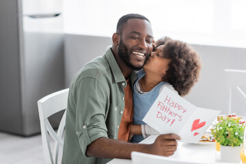 curly african american girl kissing cheek of happy father with greeting card on fathers day.