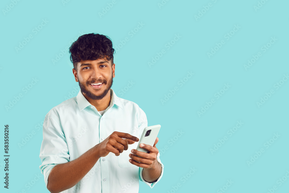 Young satisfied Indian man smiling holding phone and looking at camera typing SMS message in application and chatting with friends via messenger dressed in white shirt stands on turquoise background Young satisfied Indian man smiling holding phone and looking at camera typing SMS message in application and chatting with friends via messenger dressed in white shirt stands on turquoise background