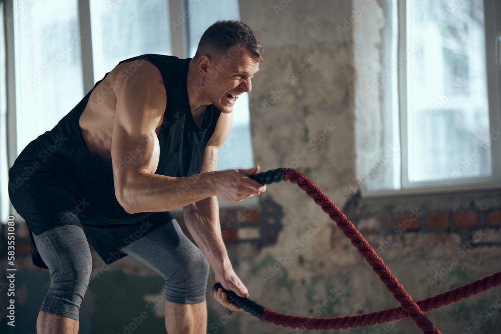 Obraz premium Coach. Young man with sportive, strong, muscular body keeping body toned, training with sports equipment, pulling battle ropes indoors. Concept of sportive lifestyle, body care, fitness, hobby, health