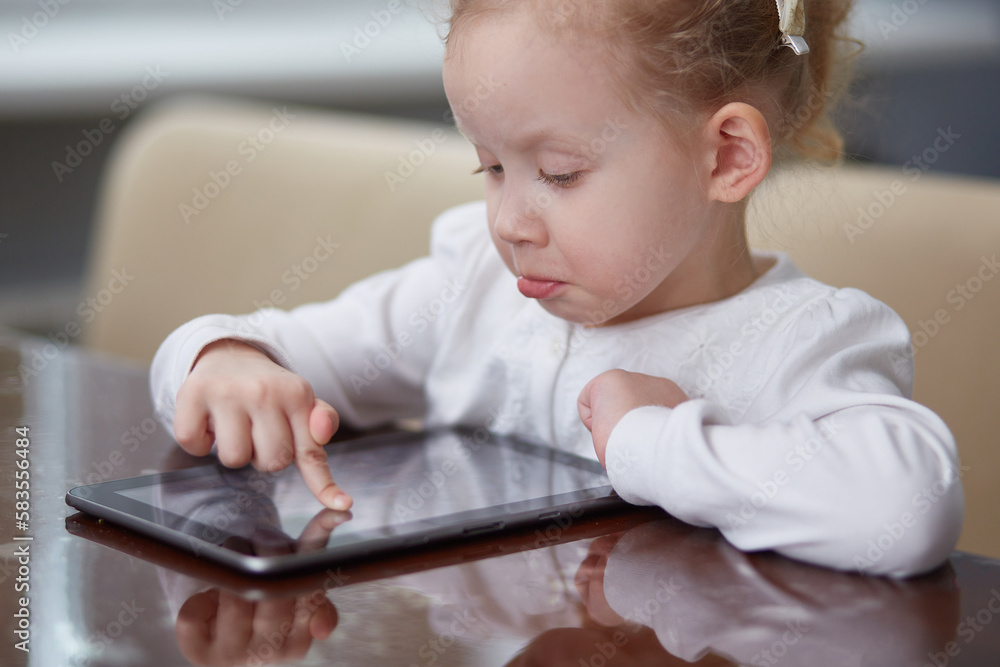 A little surprised girl is playing with a digital wireless tablet computer at home. The concept of a child and electronic devices. The concept of education and learning ability. A little surprised girl is playing with a digital wireless tablet computer at home. The concept of a child and electronic devices. The concept of education and learning ability.