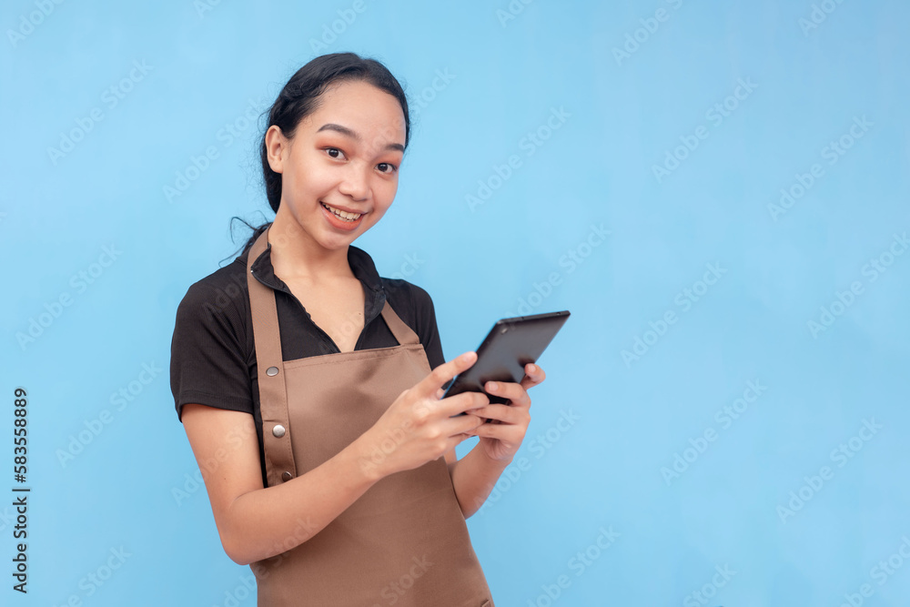A young asian female waitress or barista taking an order on her tablet while smiling. Wearing a brown apron and black shirt welcoming a customer and against a light blue background. A young asian female waitress or barista taking an order on her tablet while smiling. Wearing a brown apron and black shirt welcoming a customer and against a light blue background.