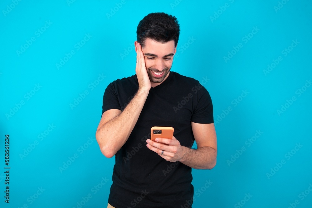 Young man wearing black T-shirt over blue studio background holding in hands cell reading sms using new app 5g Young man wearing black T-shirt over blue studio background holding in hands cell reading sms using new app 5g