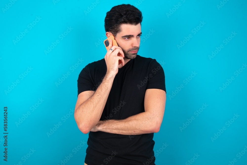 Sad Young man wearing black T-shirt over blue studio background talking on smartphone. Communication concept. Sad Young man wearing black T-shirt over blue studio background talking on smartphone. Communication concept.