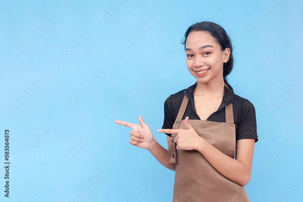 A young female barista of asian descent wearing a brown apron and black shirt finger pointing something to the left. Against a light blue background. A young female barista of asian descent wearing a brown apron and black shirt finger pointing something to the left. Against a light blue background.