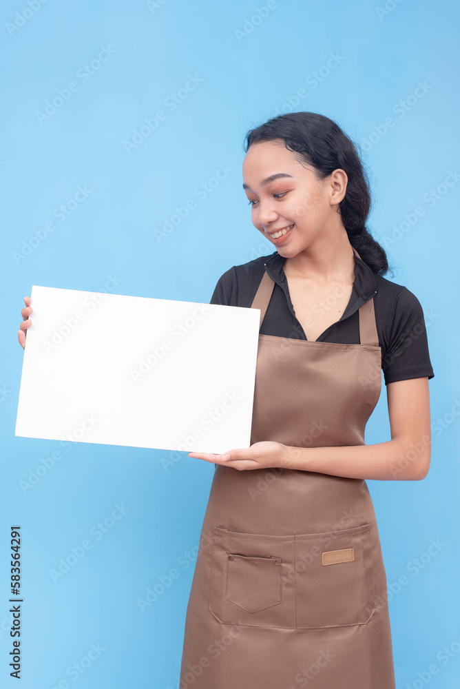 A young asian female waitress or barista holding a blank white signage. Wearing a brown apron and black shirt welcoming a customer and against a light blue background. A young asian female waitress or barista holding a blank white signage. Wearing a brown apron and black shirt welcoming a customer and against a light blue background.
