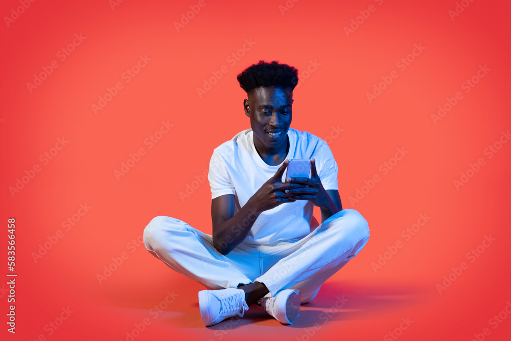 Cheerful black man using smartphone, sitting on floor Cheerful black man using smartphone, sitting on floor