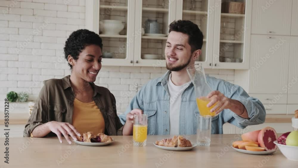 Healthy food concept. Happy international couple pour each other freshly squeezed orange juice and smile while sitting at home in the kitchen Healthy food concept. Happy international couple pour each other freshly squeezed orange juice and smile while sitting at home in the kitchen
