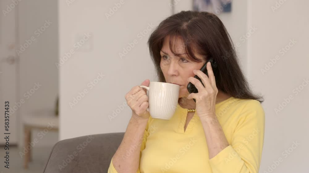 Cheerful woman enjoying drinking coffee, smiling talking on mobile cell phone sitting in home chair Cheerful woman enjoying drinking coffee, smiling talking on mobile cell phone sitting in home chair