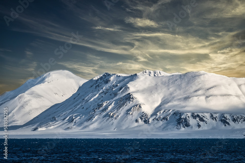 2022-05-09 SNOW COVERED SHARP PEAKS WITH SOME ROCK EXPOSED IN THE ARCTIC WITH A LIGHTY CLOUDY SKY NEAR SVALBARD NORWAY