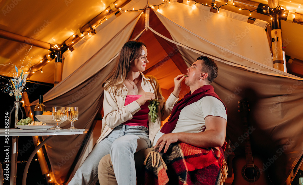 Happy lovely couple relaxing in glamping on summer evening and eating grapes near cozy bonfire. Luxury camping tent for outdoor recreation and recreation. Lifestyle concept