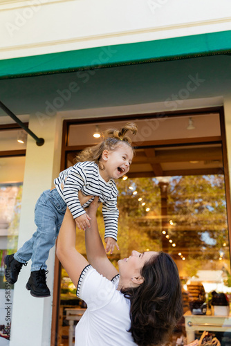 cheerful mother lifting happy baby girl near showcase of shop in Miami.