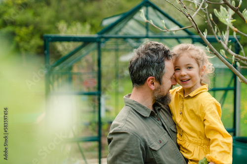Father with his little daughter laughing in front of eco greenhouse, sustainable lifestyle.