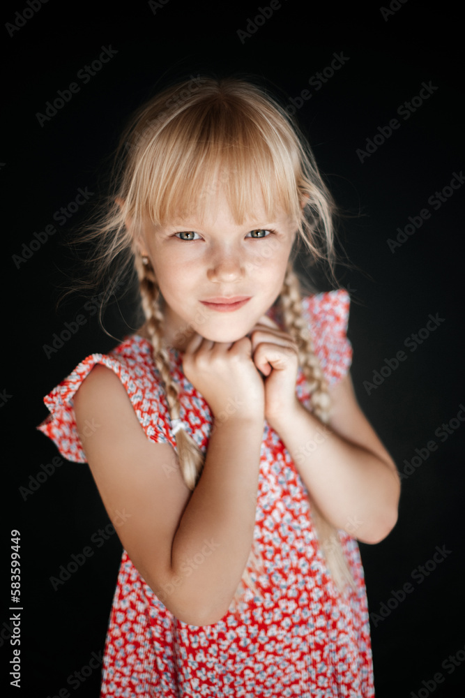 Portrait of an 8-year-old blonde girl with two pigtails, in a summer floral dress on a dark background, smiling and looking at the camera. Portrait of an 8-year-old blonde girl with two pigtails, in a summer floral dress on a dark background, smiling and looking at the camera.