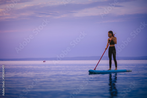 Young beautiful girl surfer paddling on surfboard on the lake at sunrise