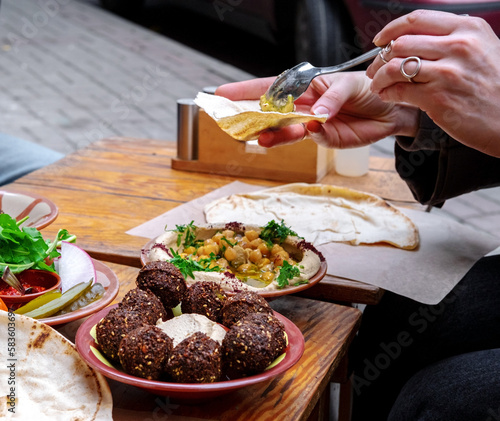 Close-up of women's hands holding bread and spreading hummus. A plate of falafel and vegetables on the table. Middle East cuisine.