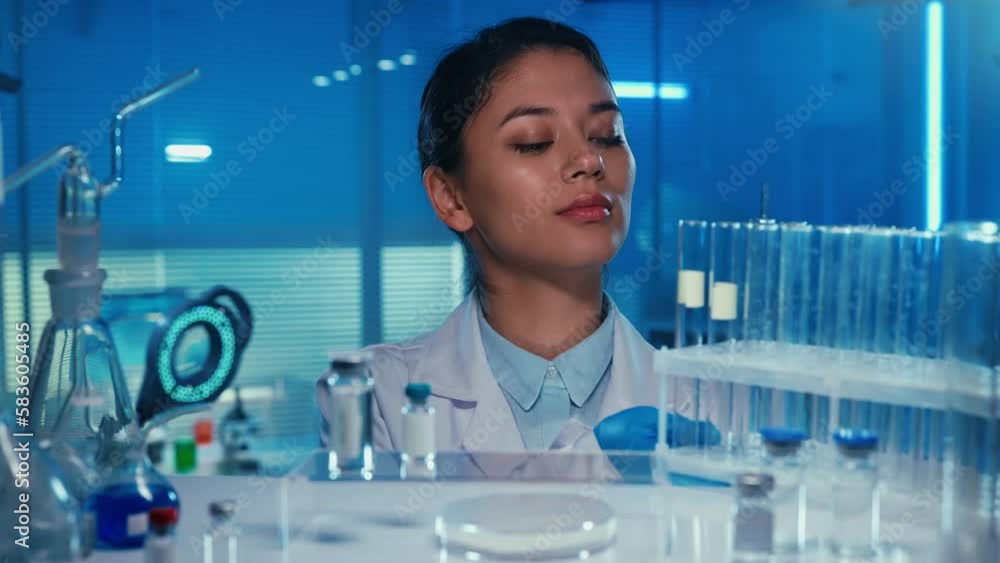 An Asian woman doctor puts a stand with test tubes on the shelf of a medical cabinet. Korean female laboratory assistant in a white medical coat in a laboratory or hospital with blue light. Close up. An Asian woman doctor puts a stand with test tubes on the shelf of a medical cabinet. Korean female laboratory assistant in a white medical coat in a laboratory or hospital with blue light. Close up.