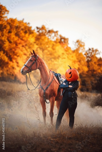 Vertical shot of a female with pumpkin head standing next to a horse in autumn