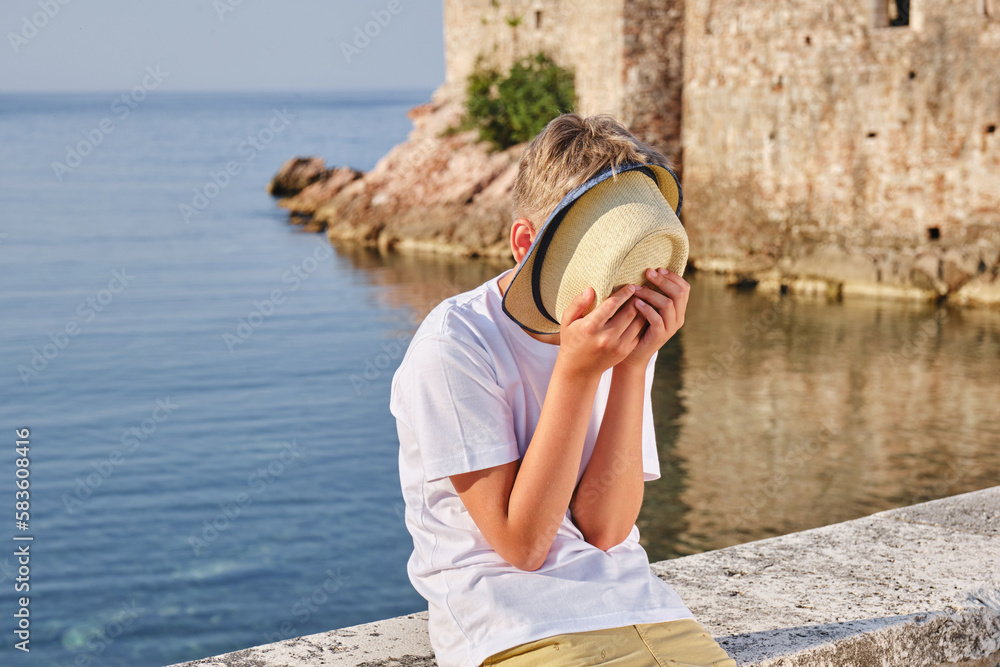 A teenage boy in a white T-shirt sits near the sea and covers his face with a straw hat.