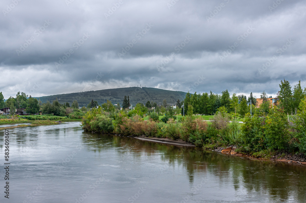 Fairbanks, Alaska, USA - July 27, 2011: Standing on Cushman bridge over Chena River, looking east landscape with green Griffin park, dark green hill under gray cloudscape