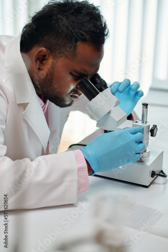 Side view of young African American male scientist looking in microscope while sitting in laboratory and studying new chemical substances