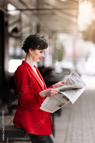 Stylish beautiful brunette woman wearing red jacket holds newspaper and reads news in the morning outdoors by the office centre. Elegant businesswoman has break for new information