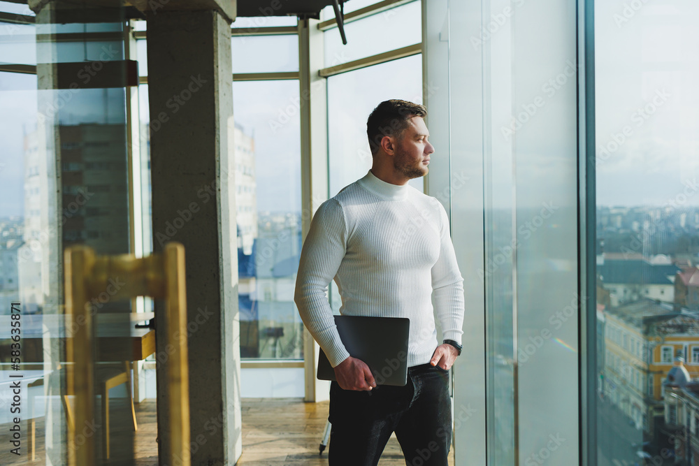 A young man in a modern workspace stands near a large window. A man in casual clothes with a laptop in his hands. Remote work. Nice young manager A young man in a modern workspace stands near a large window. A man in casual clothes with a laptop in his hands. Remote work. Nice young manager