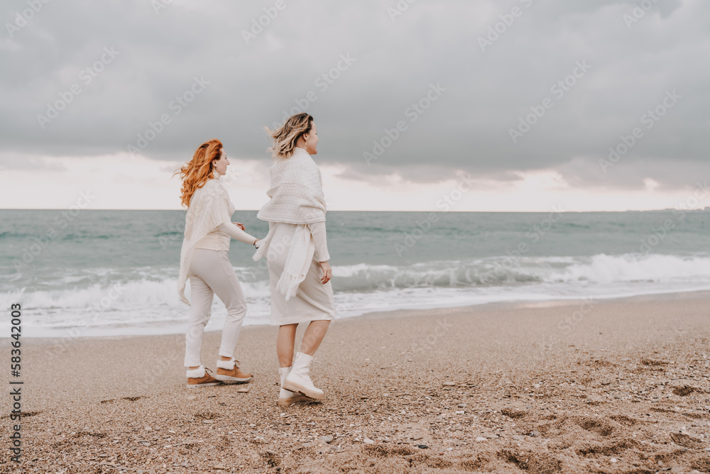 Women sea walk friendship spring. Two girlfriends, redhead and blonde, middle-aged walk along the sandy beach of the sea, dressed in white clothes. Against the backdrop of a cloudy sky and the winter Women sea walk friendship spring. Two girlfriends, redhead and blonde, middle-aged walk along the sandy beach of the sea, dressed in white clothes. Against the backdrop of a cloudy sky and the winter
