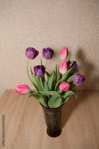 Fototapeta Naklejka Na Ścianę i Meble -  A bouquet of beautiful purple and pink tulips stand in a glass vase on a wooden table, against the background of a textured wall. Image for your creative design or illustrations.