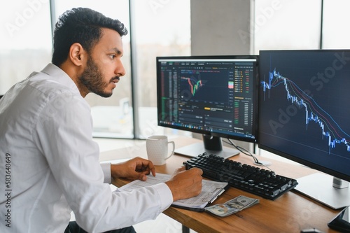 Side view of smart Indian crypto trader, investor, analyst broker, using laptop and smartphone analyzing digital cryptocurrency exchange, stock market charts, thinking of investing and funds risks.