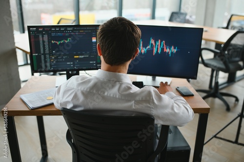 Man trader in formalwear sitting at desk in frot of monitors with charts and data at office browsing laptop checking documents analyzing stocks price changes concentrated. photo