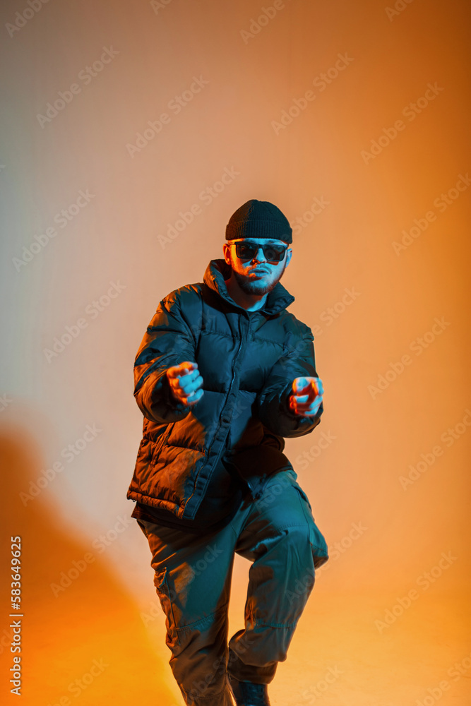 Cool trendy stylish man dancer with fashion sunglasses in trendy black clothes with a hat and a down jacket is dancing in a creative color studio with warm yellow and cold blue light