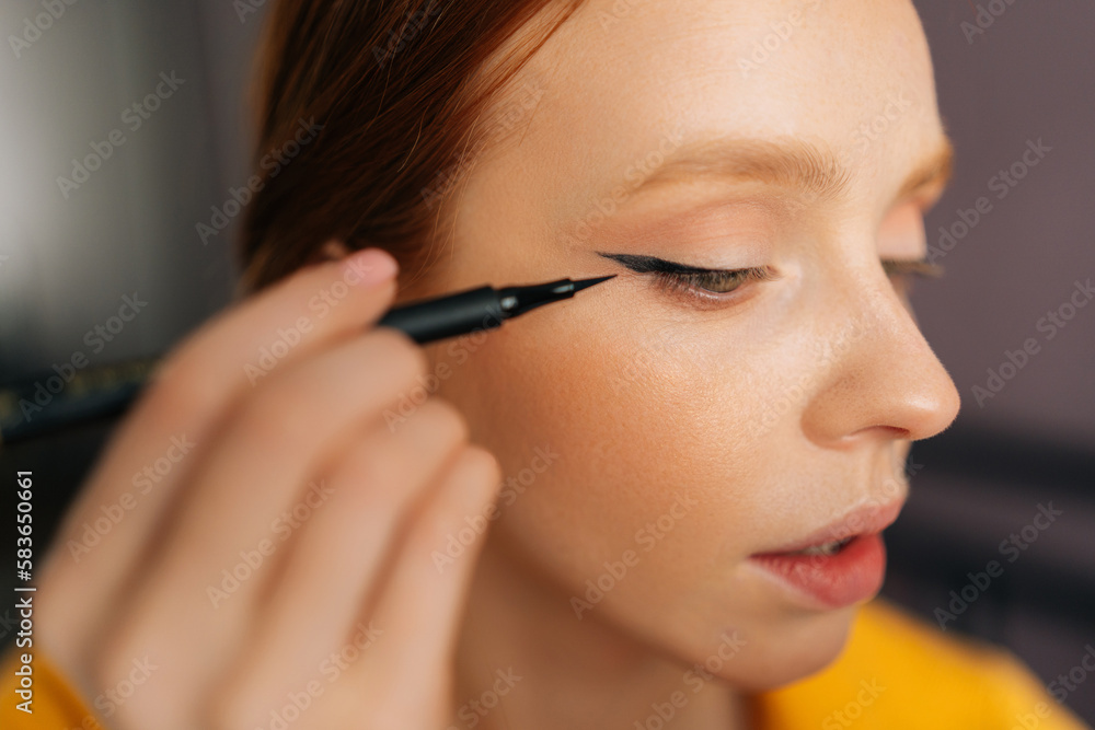 Closeup face of attractive young woman using contour brush drawing arrows on eyelids looking at reflection in mirror. Beautiful redhead female applying eyeliner drawing cat eyes makeup sitting on bed.