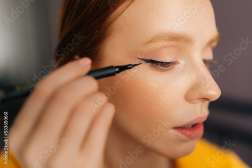 Closeup face of attractive young woman using contour brush drawing arrows on eyelids looking at reflection in mirror. Beautiful redhead female applying eyeliner drawing cat eyes makeup sitting on bed. © dikushin